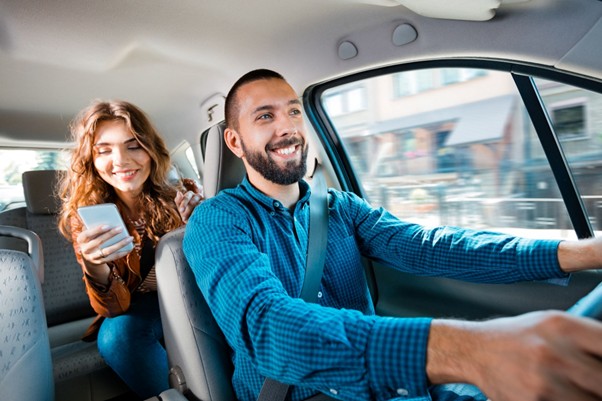 Smiling e-hailing driver in a blue shirt driving a passenger using a ride-share app, illustrating the need for e-hailing insurance in South Africa.