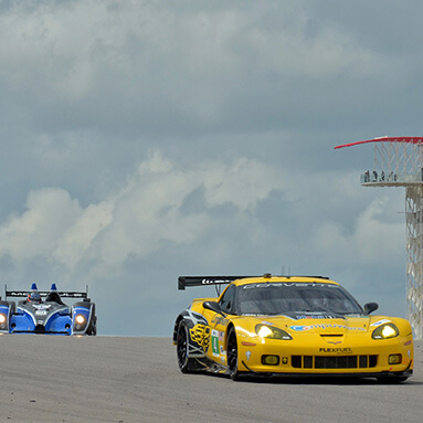Side front view of a yellow Chevrolet Corvette racing car on the race course with cloudy sky in the background.