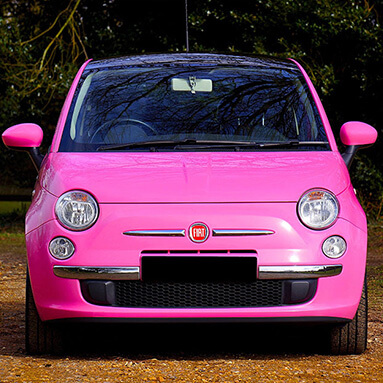 Parked pink Fiat 500, front view, on the grass with trees in the background.