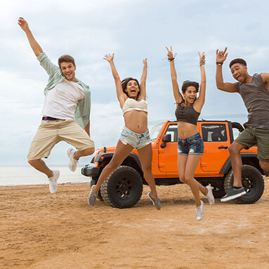 Group of friends jumping up for joy, at the beach with an orange jeep in the background.