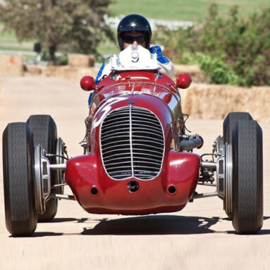 Maroon classic maserati being driven by passenger wearing a helmet and sunglasses.