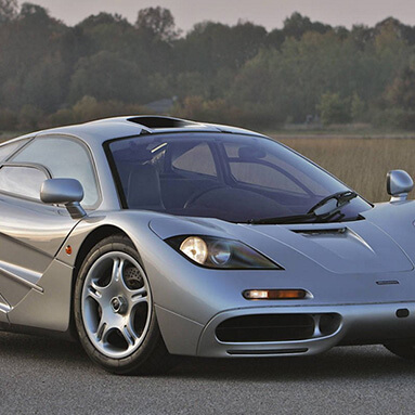 Front side view of a grey McLaren F1 on the road with trees in the background. 