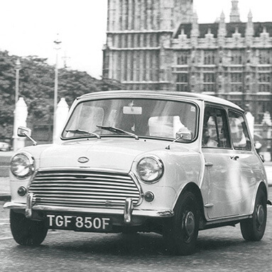 White vintage Mini Cooper on the road with British castle in the background.
