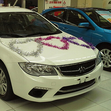 Side front view of a White Mitsubishi Lancer decorated with tinsel on the bonnet.