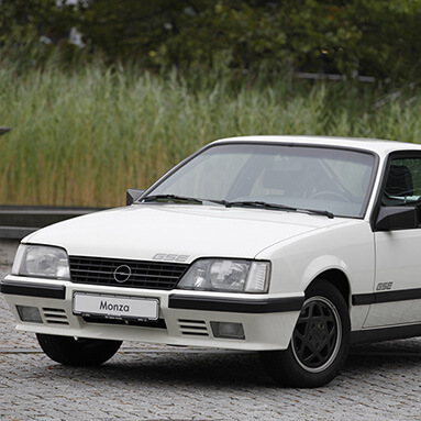 Classic white opel monza, parked sideways in parking lot, with green reeds in background.