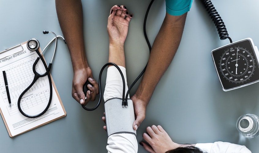 Doctor checking patient’s blood pressure, with stethoscope, clipboard and pen on table.