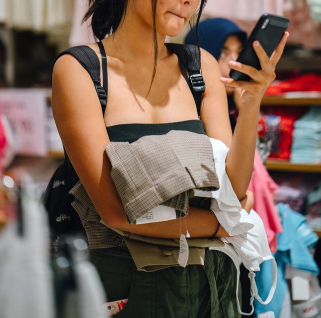 Young woman shopping, holding clothes while looking at her phone.