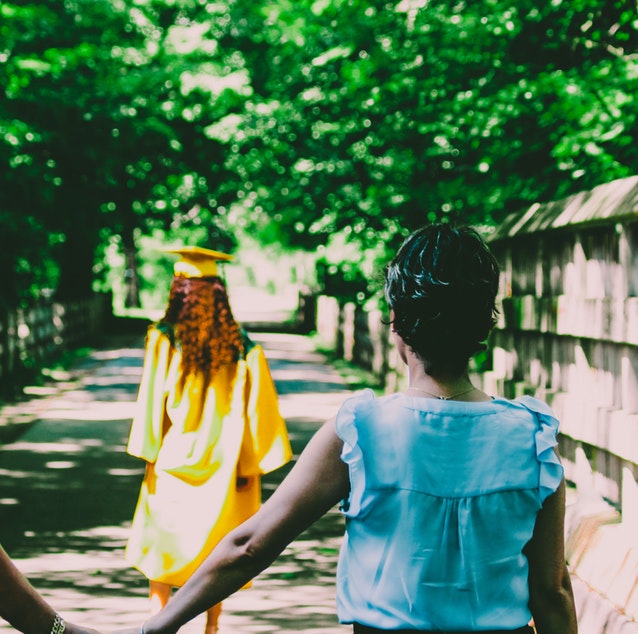 Parents holding hands watching their daughter wearing a graduation gown and walking over a bridge.