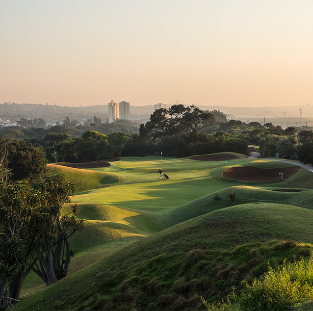 Durban Country Club green rolling hills on golf course, two men playing in the middle and city buildings in background.