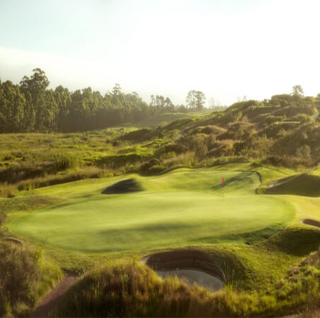 Fancourt Links Golf Course hole with red flag, surrounded by trees on the left and small sand dunes and hills on right.