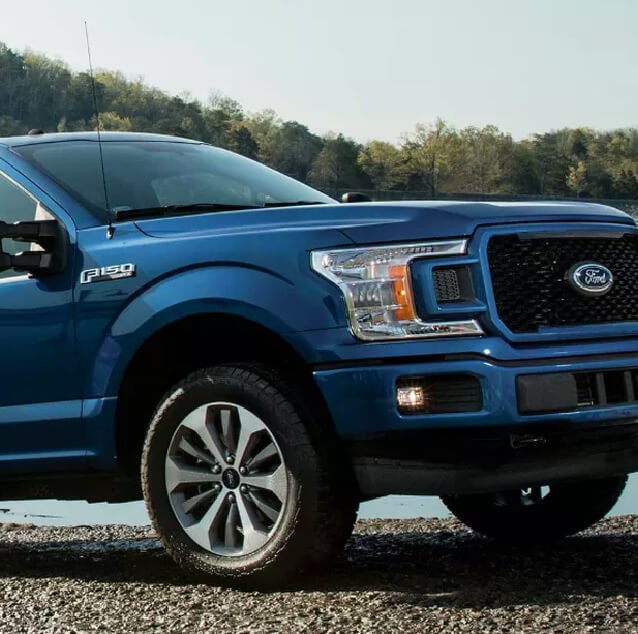 Front side view of a blue Ford F Series with a bush in the background.