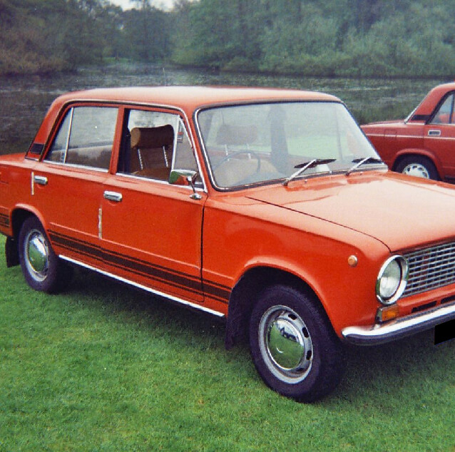 Front side view of a red Lada 1200 with a green bush in the background.