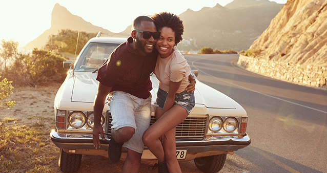 A man and woman posing in front of a vintage car on a mountain pass