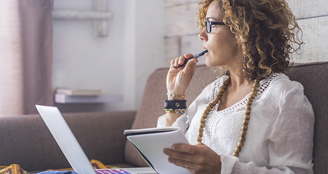 A pondering woman sitting on a couch with a laptop and notebook