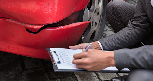 A person filling out a form on a clipboard next to a dented car bumper