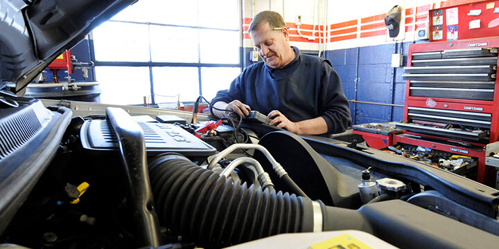 Open car hood/bonnet with mechanic checking the car’s battery in the background