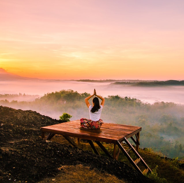 Woman sitting on top of wooden platform with her hands over her head and palms together, overlooking a cloudy, scenic view of clouds, tree tops and an orange sunset.