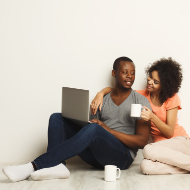 Couple having a warm beverage looking at each other with laptop on man's lap as they sit on the floor | Our name says it all | Hippo.co.za partner
