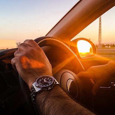 Mans hands on steering wheel with sun reflecting in side mirror as he drives on the road | History of Dialdirect | Proud hippo partner