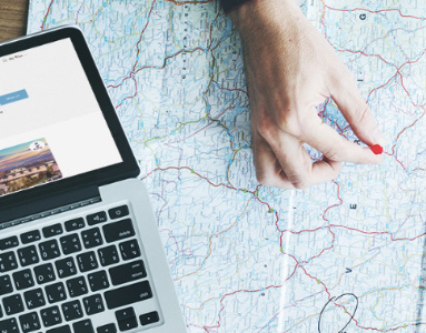 Laptop screen open to display Travelstart page with a hand holding a pin on a map to indicate a destination the person would like to travel to | What we offer | Hippo.co.za travel partner