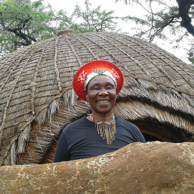  Smiling Zulu lady standing in front of a traditional Zulu hut.