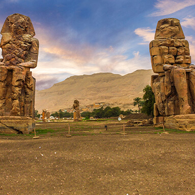 Brown mountain rock face in between Egyptian statues and green land, and trees in between.