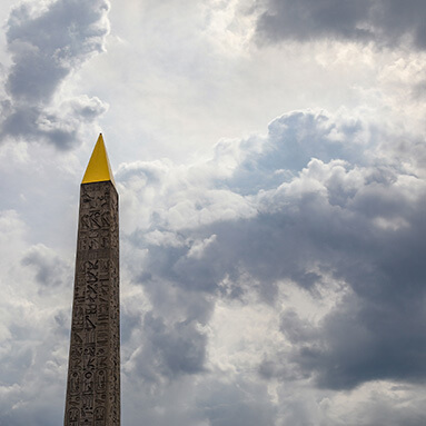 Grey skies hovering over a tall heritage building, that has a golden triangular tip and hieroglyphics.