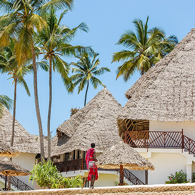 Maasai tribesman looking up at white thatched roofing accommodation, with palm trees and blue skies.