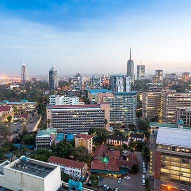 Topographical view of buzzing, urban Nairobi buildings, with a striking blue sky, and semi-lit businesses.