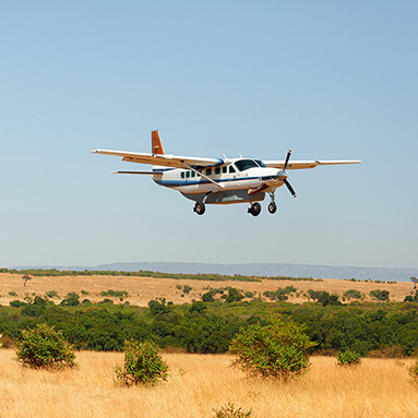 White private aeroplane flying over green and brown bush with small green trees and blue skies.