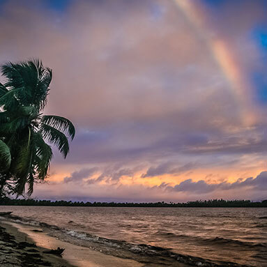 View from shoreline of a rainbow over the sea at dusk.