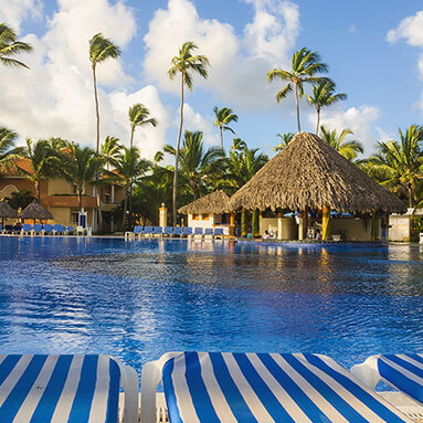 Blue and white pool loungers overlooking a big swimming pool with thatched gazebos and trees in the background.