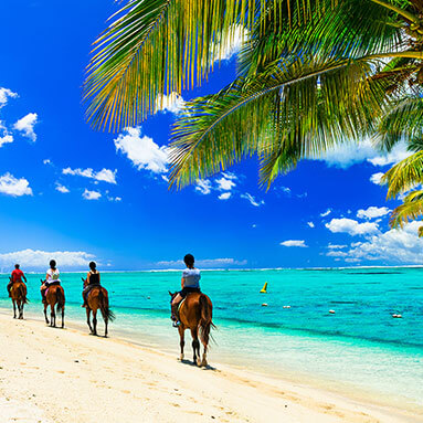 People on horses on the beach with blue cloudy sky and the sea in the background.