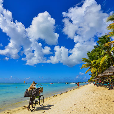 Man pushing bicycle on the beach with the sea and cloudy sky in the background.