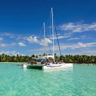 People on a boat in the sea with trees and blue cloudy sky in the background.