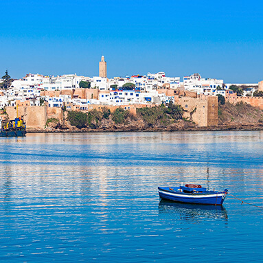 Blue boat floating in blue water, with blue and white buildings on the shore.