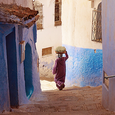 Woman carrying an item over her head, while walking through a narrow, mud corridor, with blue and white walls.