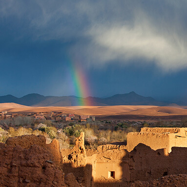 Rainbow striking through blue skies over dilapidated clay houses and mountains in distance.