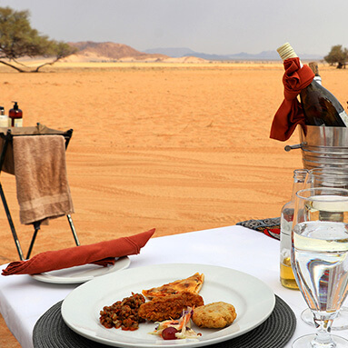 Scrumptious, light, colourful lunch on a plain white table setting in the desert with wine in a bucket and desert in the distance.