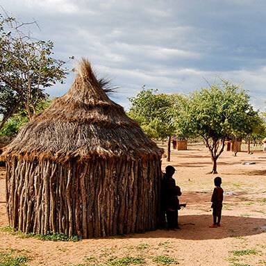 Kids outside a thatched hut with trees in the background.