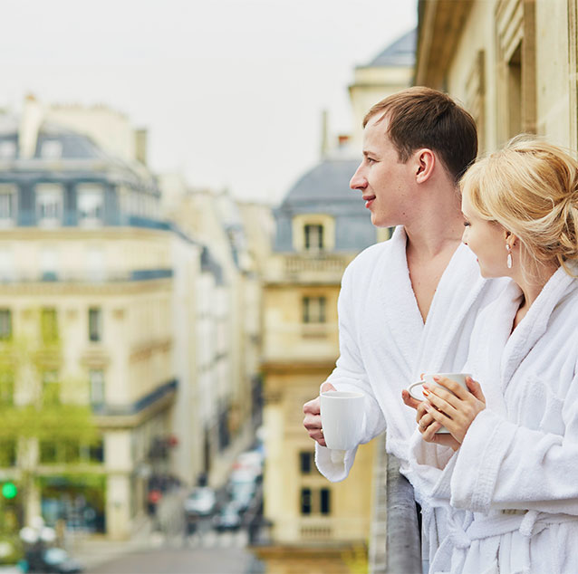 Man and woman wearing white rooms and carrying mugs standing at the balcony.