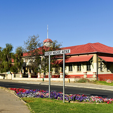 Picture of red-roofed yellow building on Robert Mugabe Avenue, with colourful flowers in the pathway.