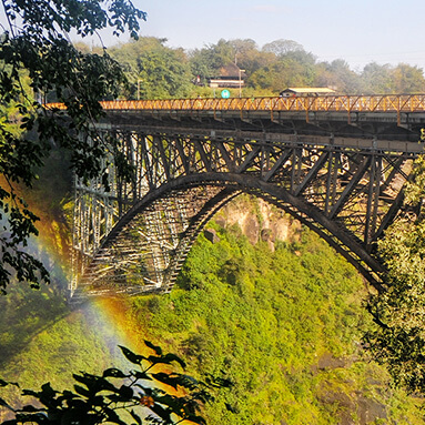 Rainbow running through Breitbach bridge on a sunny day with blue skies, luscious trees and vegetation below.