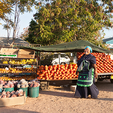 A smiling woman walking on a dirt road, across a colourful fruit and vegetable stall.