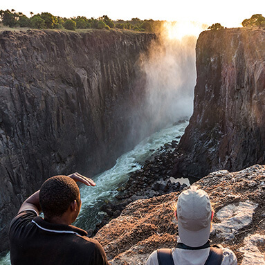 Tour guide with tourist overlooking Victoria Waterfalls, with water rushing through rocky, small river pathway.