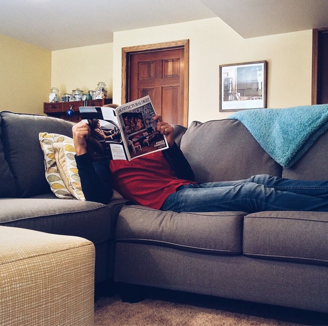 Man wearing red shirt and jeans, while reading an architecture magazine with feet up on the couch, in the living room.