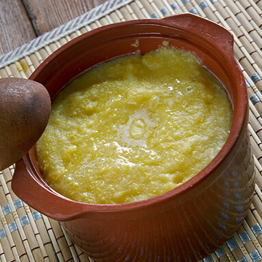 Yellow looking mashed potato traditional Botswana sweswe food in clay ceramic dish, with a wooden spoon on side.