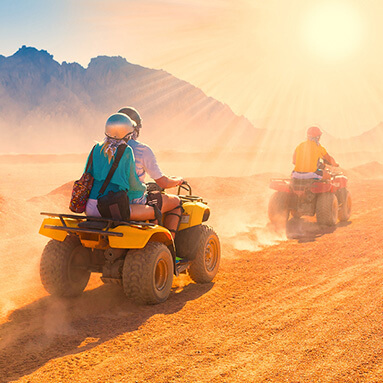 A couple, dirt biking through the desert on a sunny day with a man in front of them and dust in the air.