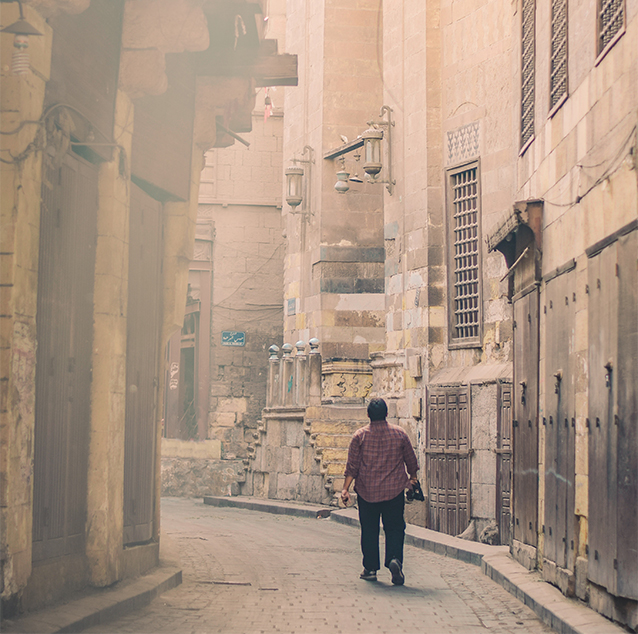 A man walking through brown high walls and apartments of Cairo City, wearing a purple shirt and holding a jacket.